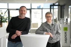 Benjamin Manzenberger und Brigitte Rothkopf von Sesotec GmbH stehen lächelnd an einem Stehtisch in einem hellen Büro- bzw. Empfangsbereich mit Sesotec-Logo im Hintergrund